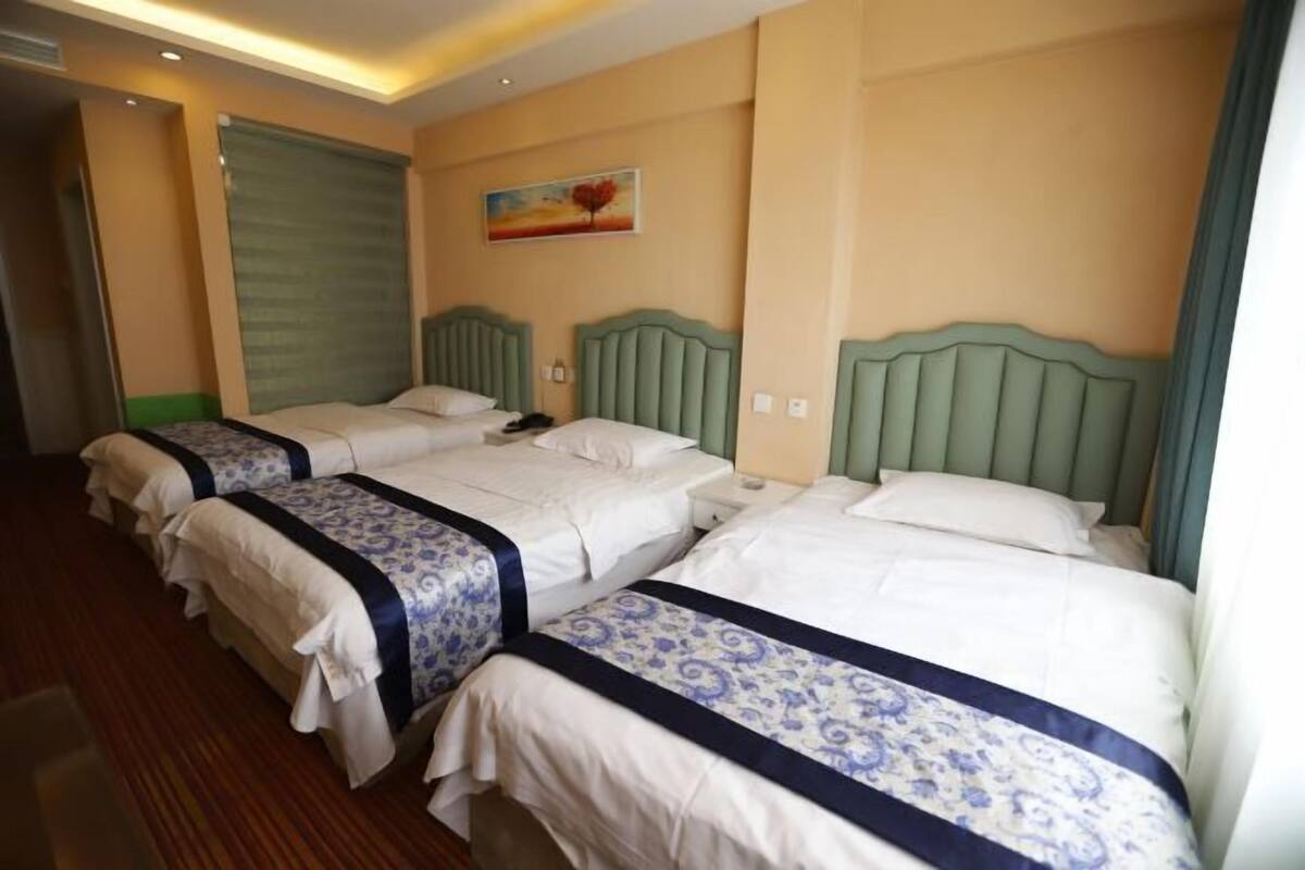 A hotel room with three beds, each with a headboard, a painting on the wall, and a closed window.