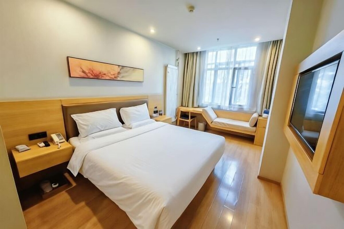 A hotel room with a large bed, a bedside table with a lamp, a TV, and a window with curtains.