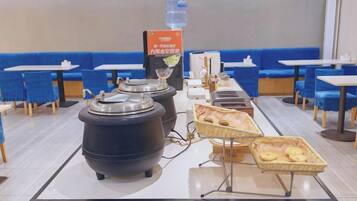 Montaje de bufé con ollas calientes, dispensador de agua y cestas de pan en un restaurante con asientos azules.
