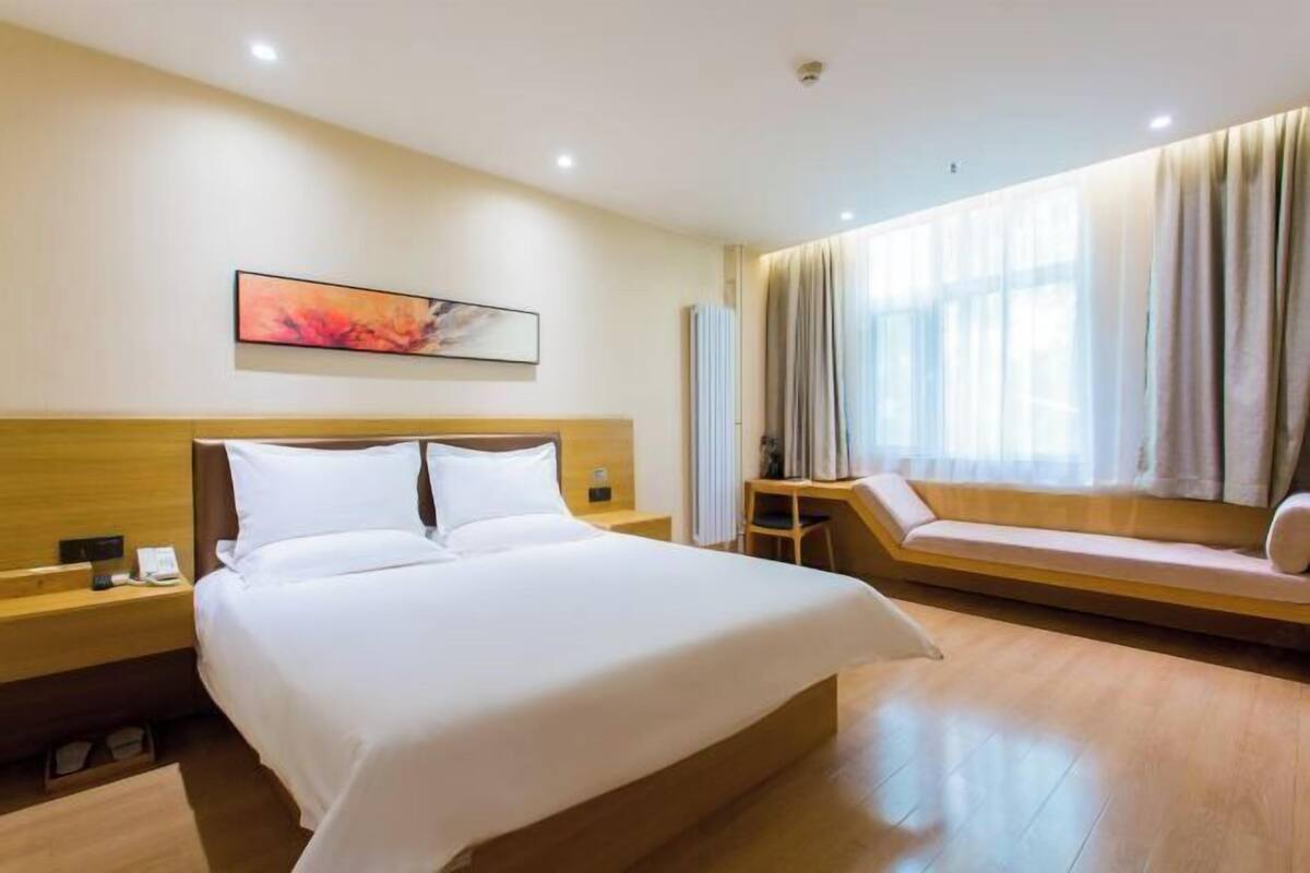 A hotel room with a large bed, a bedside table, a small table, a sofa, and a window with curtains.