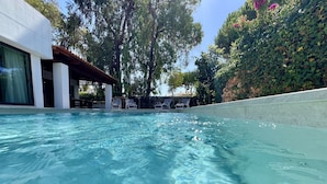 Ein klares Schwimmbecken mit Liegestühlen und einem überdachten Bereich, umgeben von viel Grün und einem Gebäude.