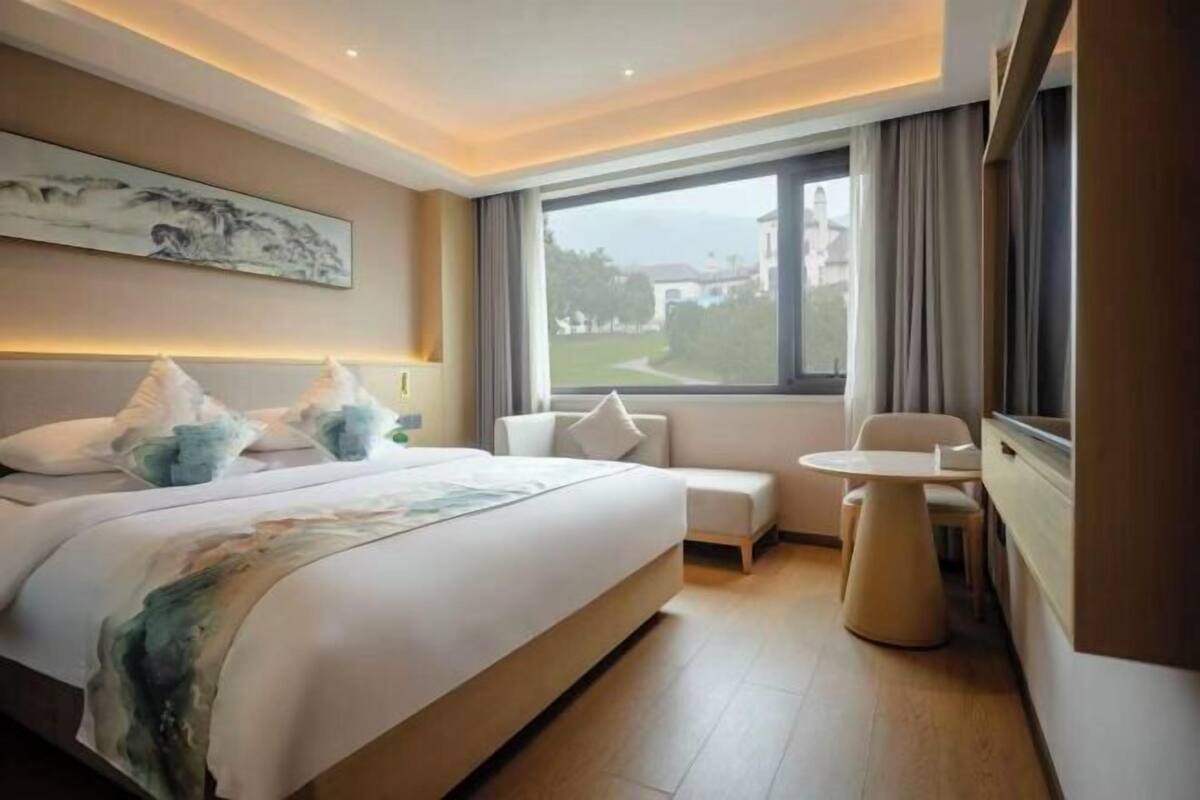 A hotel room with a bed, a small table, a chair, a window with a view, and a painting on the wall.