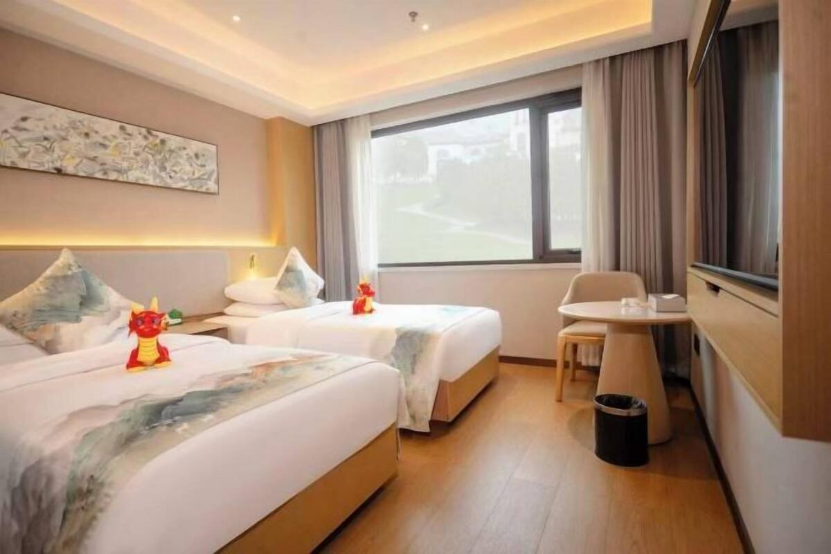 A hotel room with two beds, a small table, a chair, a TV, and a window with curtains.