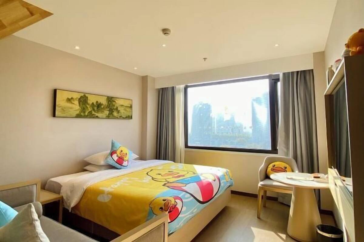 A hotel room with a bed, a chair, a small table, a window with curtains, and a painting on the wall.