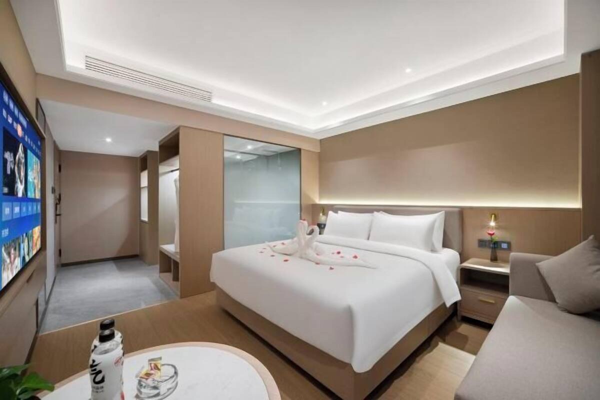 A modern hotel room with a large bed, bedside tables, a TV, and a glass partition wall.