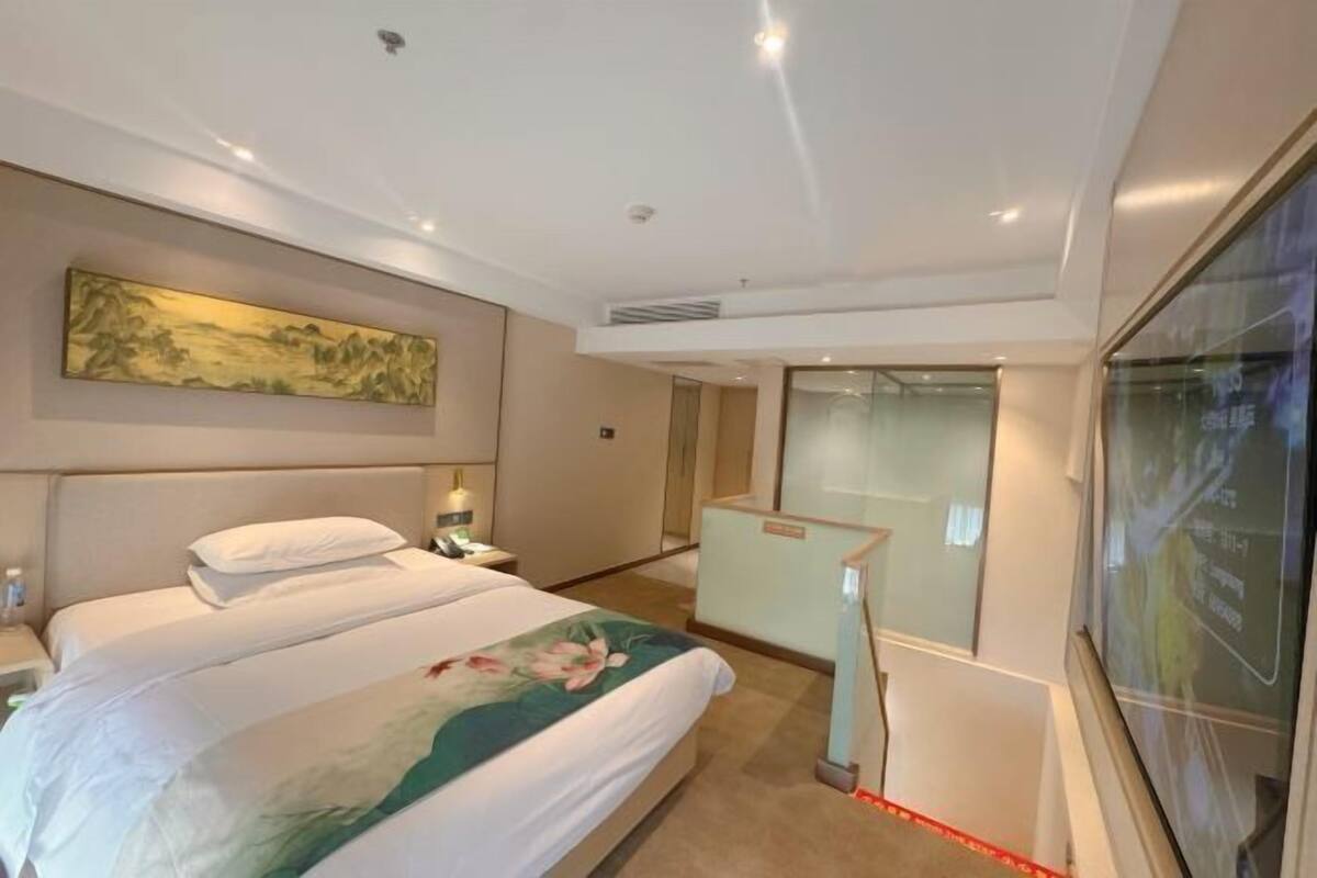 A hotel room with a bed, a nightstand, a desk, a large painting on the wall, and a glass partition.