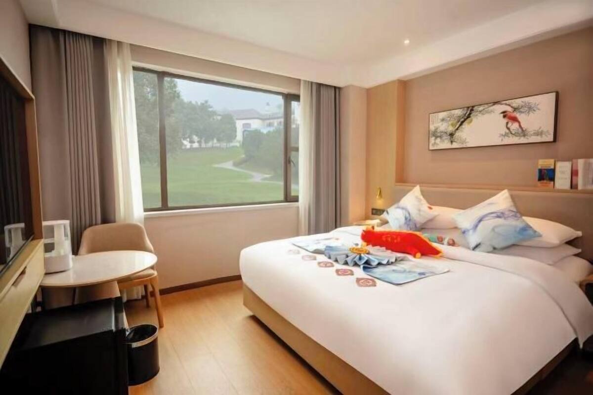 A hotel room with a bed, a small table, a chair, a window with a view of greenery, and a painting on the wall.