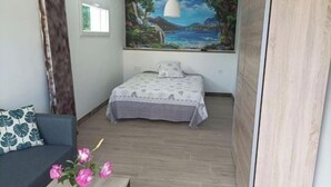 A compact room with a bed, a sofa, a small table with flowers, and a painting of a coastal landscape on the wall.
