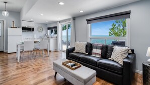 A modern living room with a black leather sofa, a dining area with white chairs, and a kitchen with white cabinets.