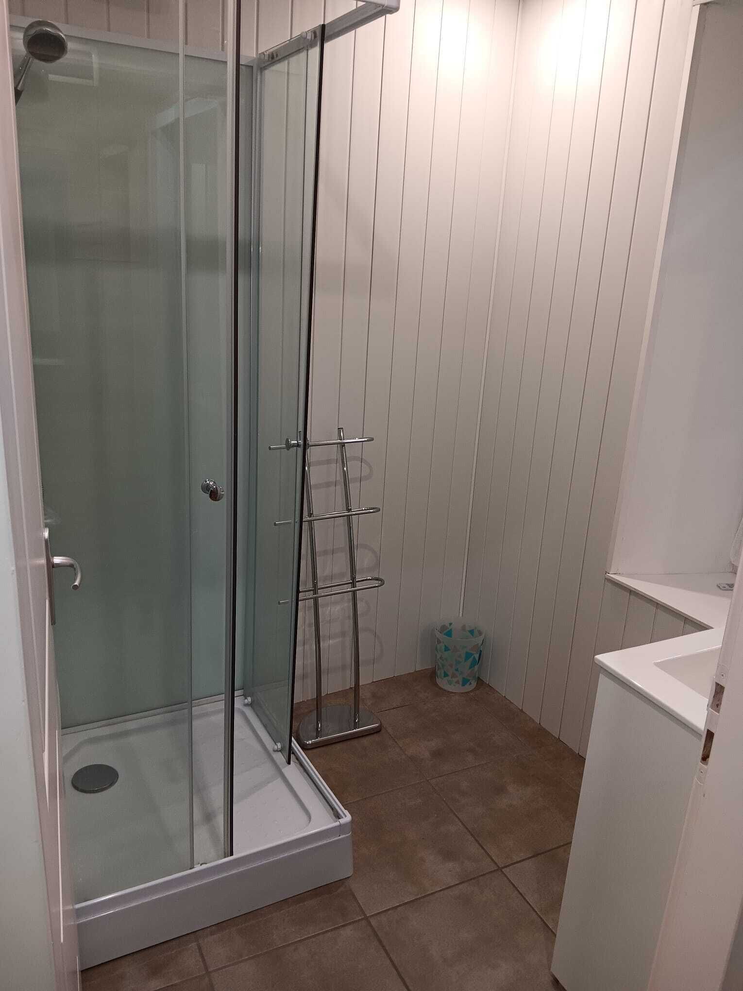 A glass-enclosed shower with a metal rack, white wooden paneling, and a tiled floor.