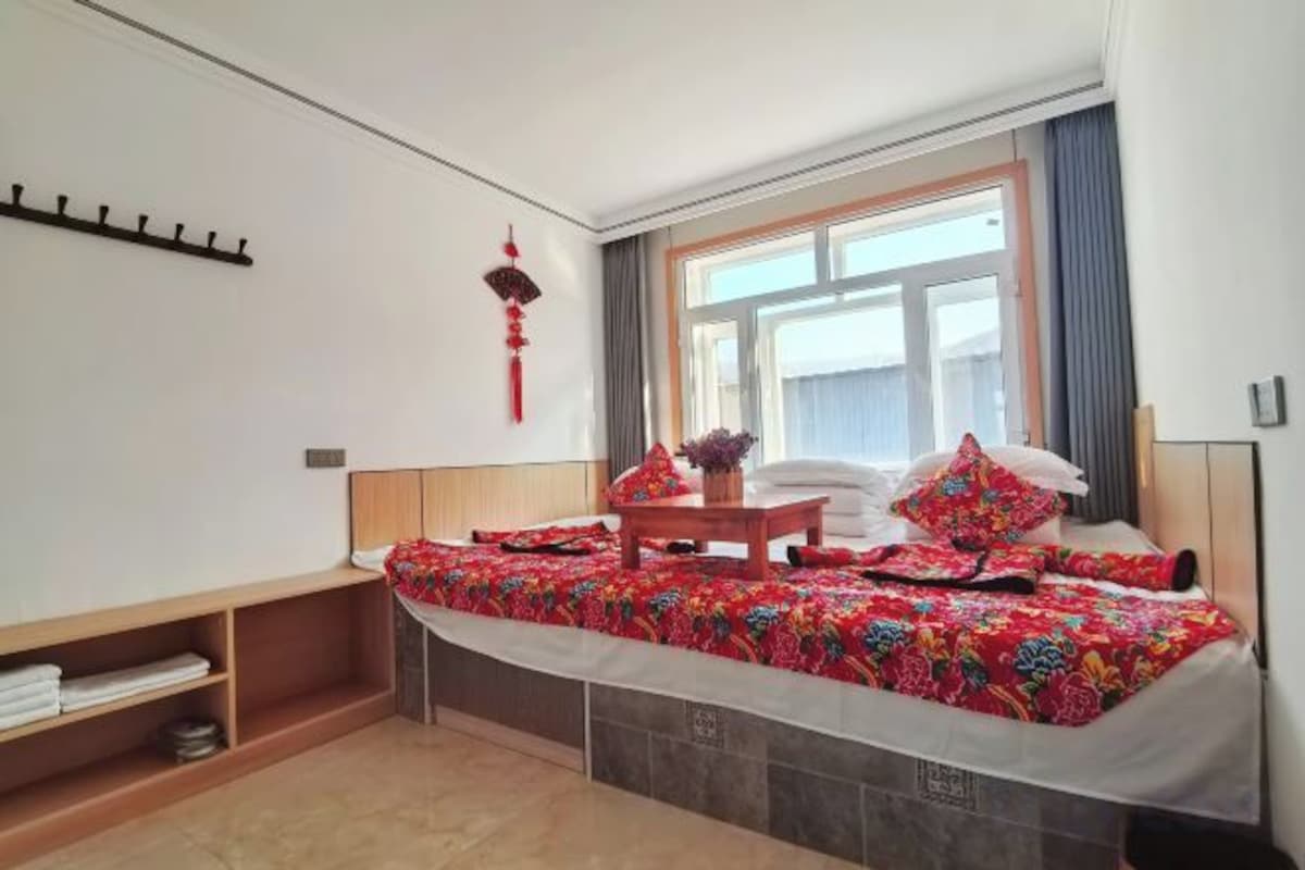 A bedroom with a bed, a small table, a window, and a wall-mounted shelf.