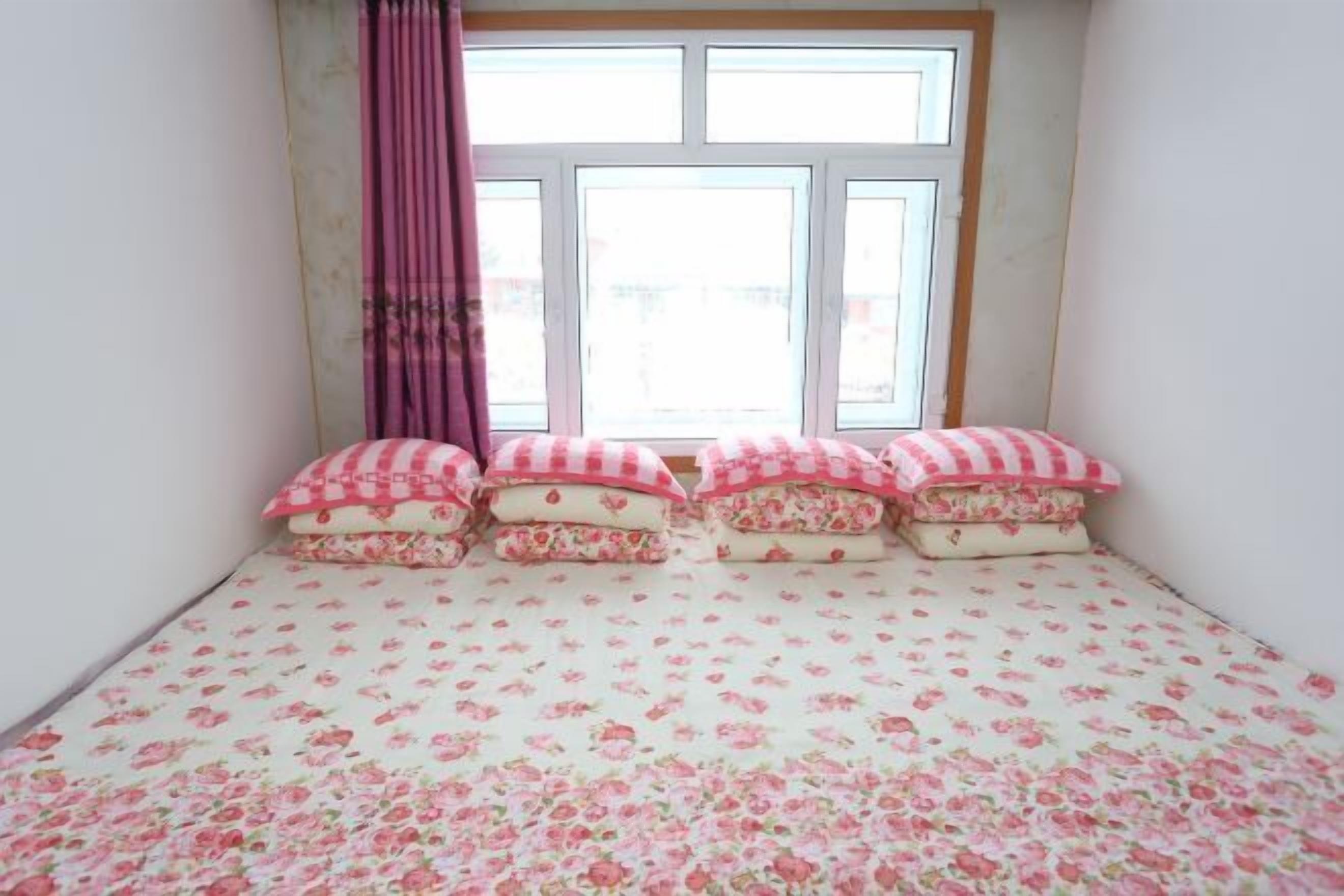 A bedroom with a floral-patterned bedspread, a window with curtains, and a view of the outside.