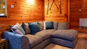Ein gemütliches Blockhaus-Interieur mit Holzbalkendecke, einem großen L-förmigen Schlafsofa mit vielen Kissen und einem Paar Ski, die an der Wand lehnen.