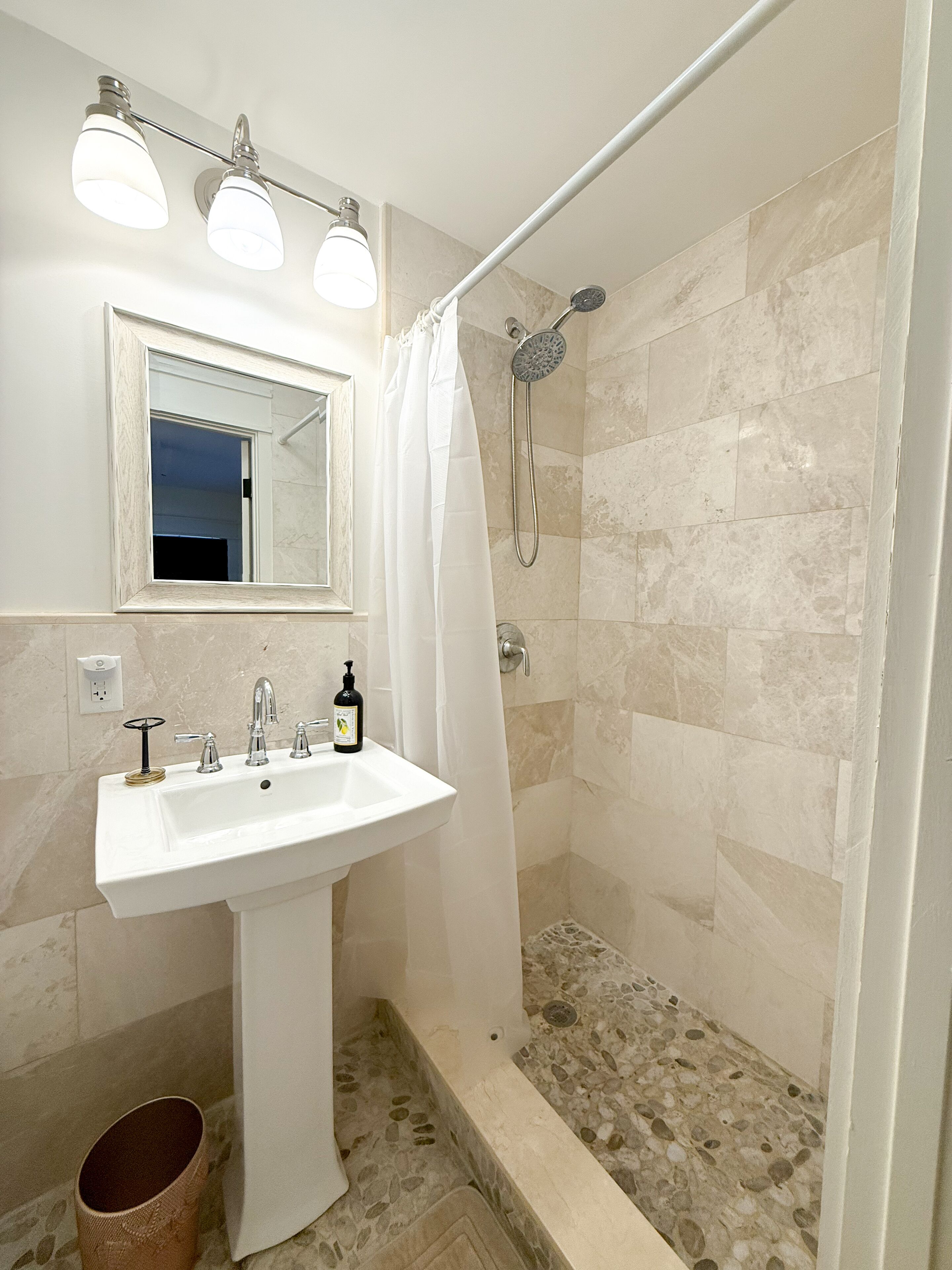 Une salle de bain avec un lavabo blanc, un miroir, une douche équipée d’un pommeau de douche amovible et un sol carrelé.