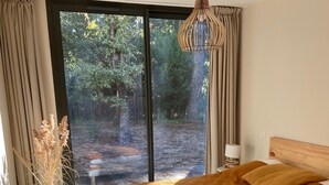 A bedroom with a large bed, a window with curtains, a hanging light fixture, and a vase with dried plants on the windowsill.
