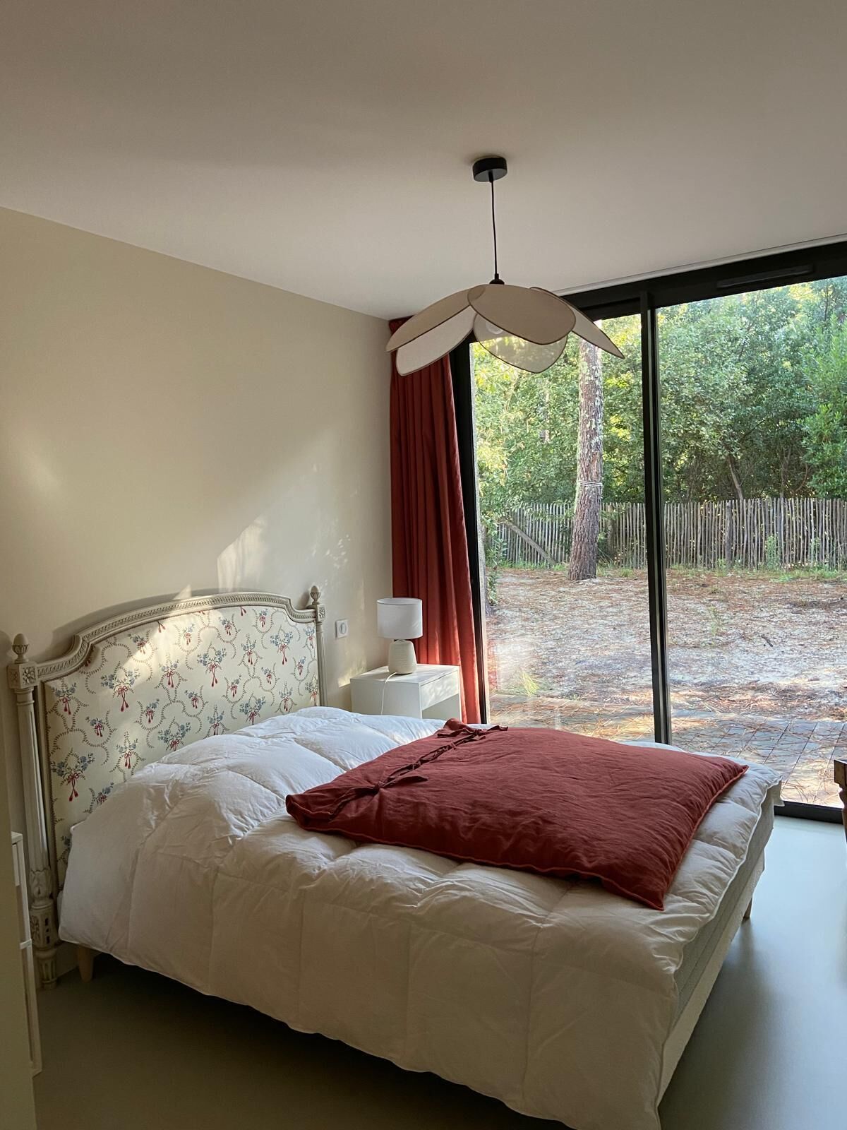 Een slaapkamer met een groot bed, een nachtkastje, een lamp en uitzicht op het groen door een raam.