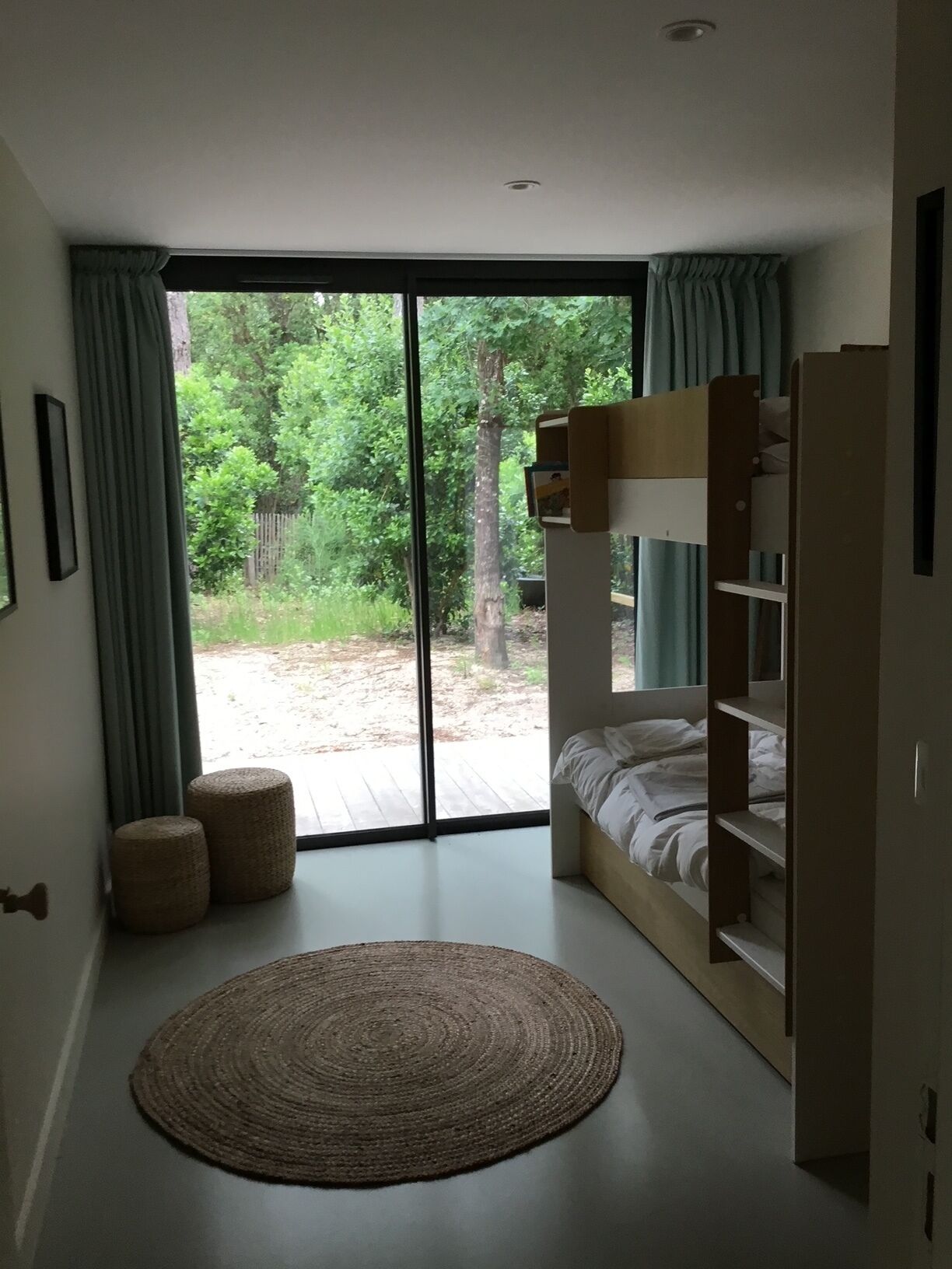 A bedroom with a bunk bed, a large window with curtains, a woven rug, and a view of a wooded area.