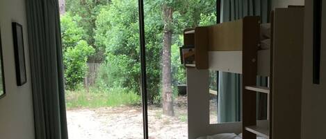 A bedroom with a bunk bed, a large window with curtains, a woven rug, and a view of a wooded area.