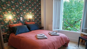 Un dormitorio con papel tapiz floral, una colcha roja, dos almohadas, una ventana con vistas a la vegetación y suelo de madera.