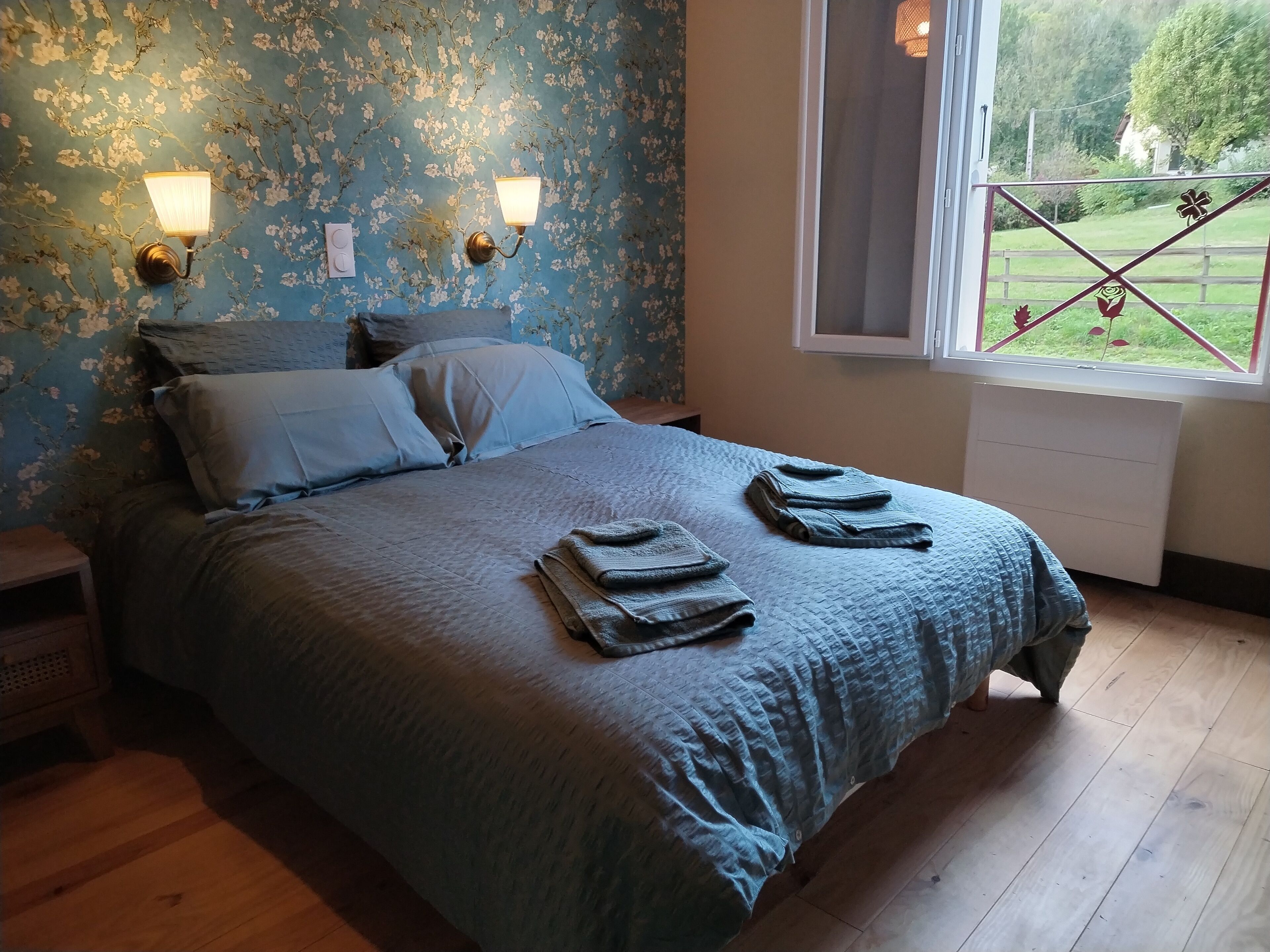 Un dormitorio con papel tapiz floral, una cama con colcha azul, dos lámparas fijadas a la pared y una ventana con vistas a un jardín.