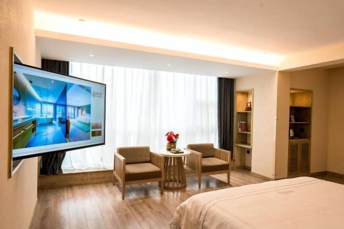 Une chambre d’hôtel comprenant un lit, une télévision, deux fauteuils, une petite table avec un vase et une armoire.