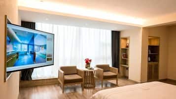 A hotel room with a bed, a TV, two armchairs, a small table with a vase, and a wardrobe.