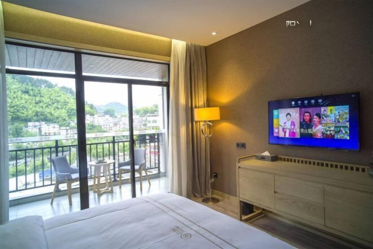 Une chambre d’hôtel avec une grande fenêtre, un lit, une télévision et un balcon équipé de chaises.