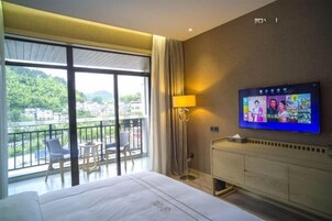 Habitación de hotel con un ventanal grande, una cama, un televisor y un balcón con sillas.