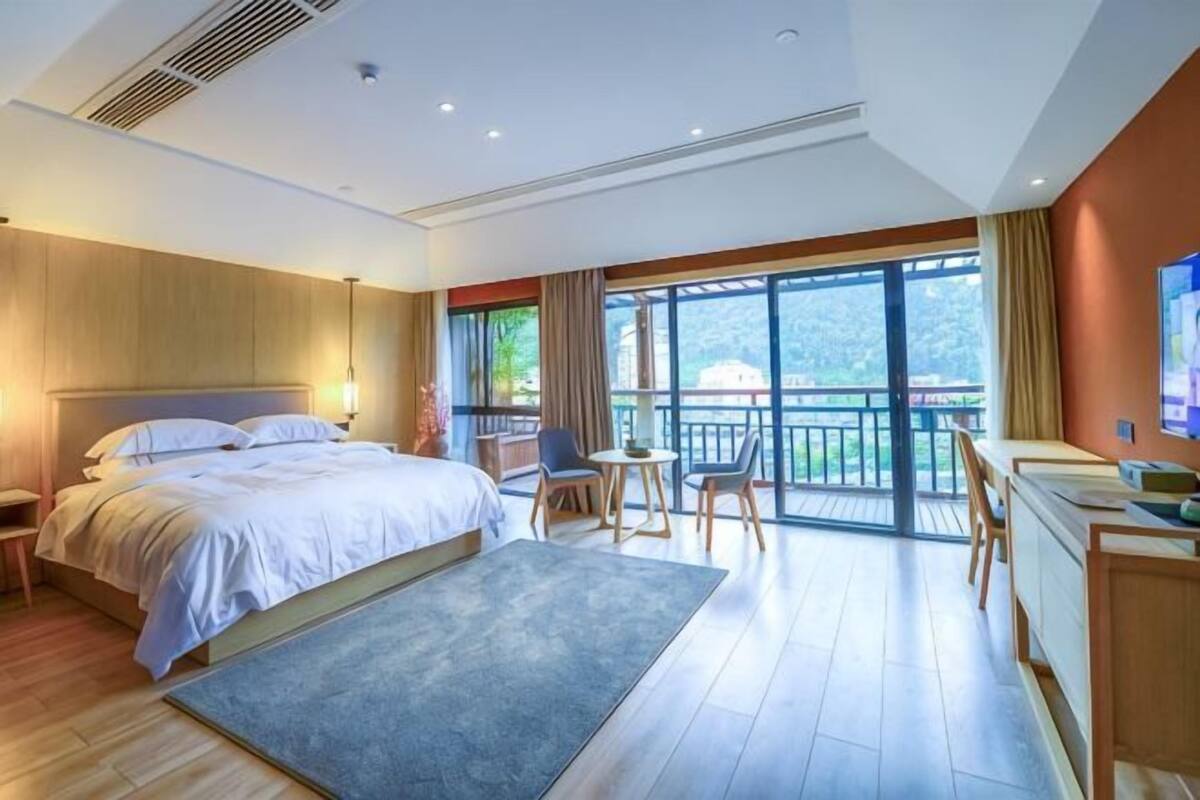 Une chambre d’hôtel avec un lit, un bureau, une chaise, un balcon et de grandes fenêtres.