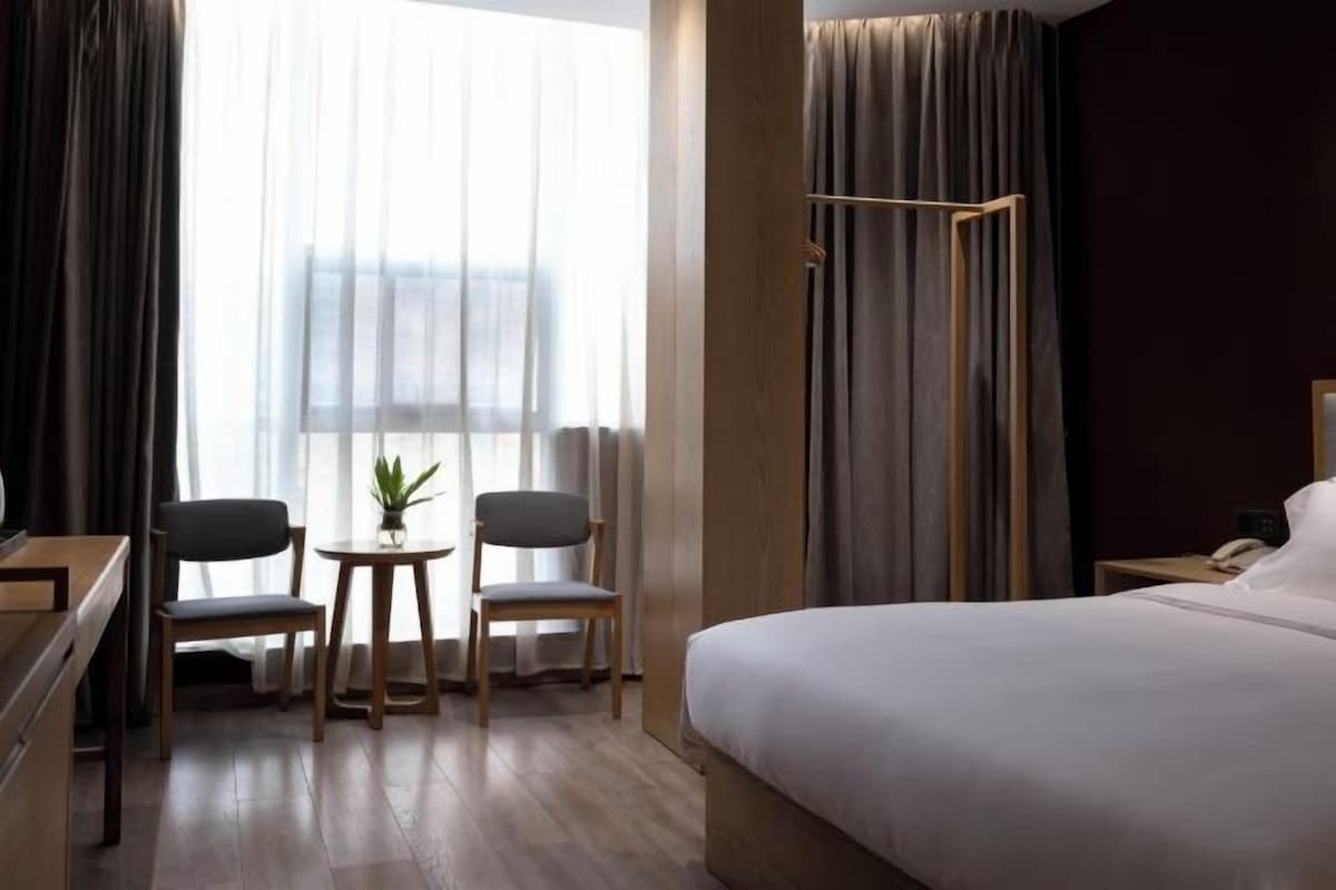 A hotel room with a bed, a desk, two chairs, a small table with a plant, and a window with curtains.
