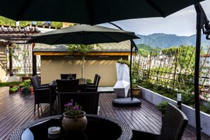 Eine Terrasse im Freien mit Tisch und Stühlen unter einem Sonnenschirm, mit Blick auf eine gebirgige Landschaft.