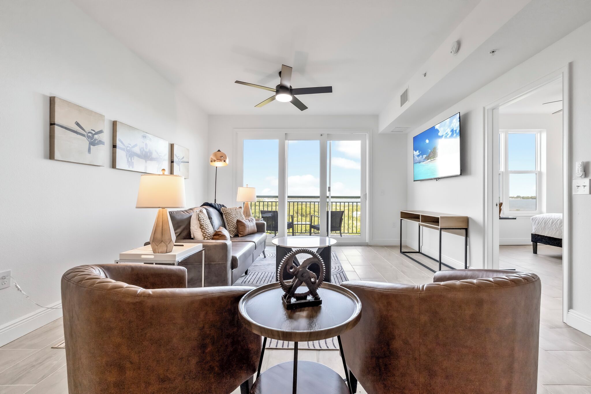 A spacious living room with a sofa, a coffee table, and a ceiling fan. There is a balcony with a view of the outdoors.