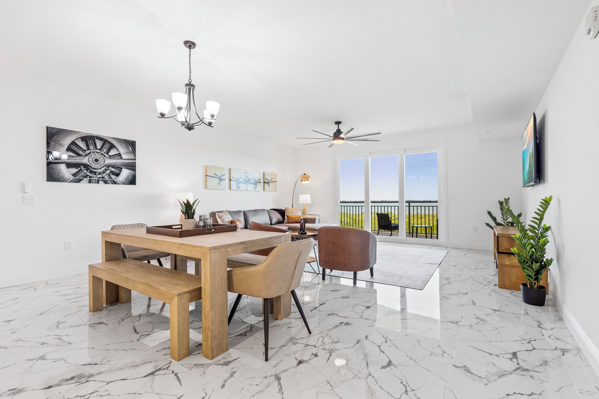 Un salon spacieux avec une table à manger en bois, des sièges confortables, un ventilateur de plafond et de grandes fenêtres offrant une vue sur l’extérieur.