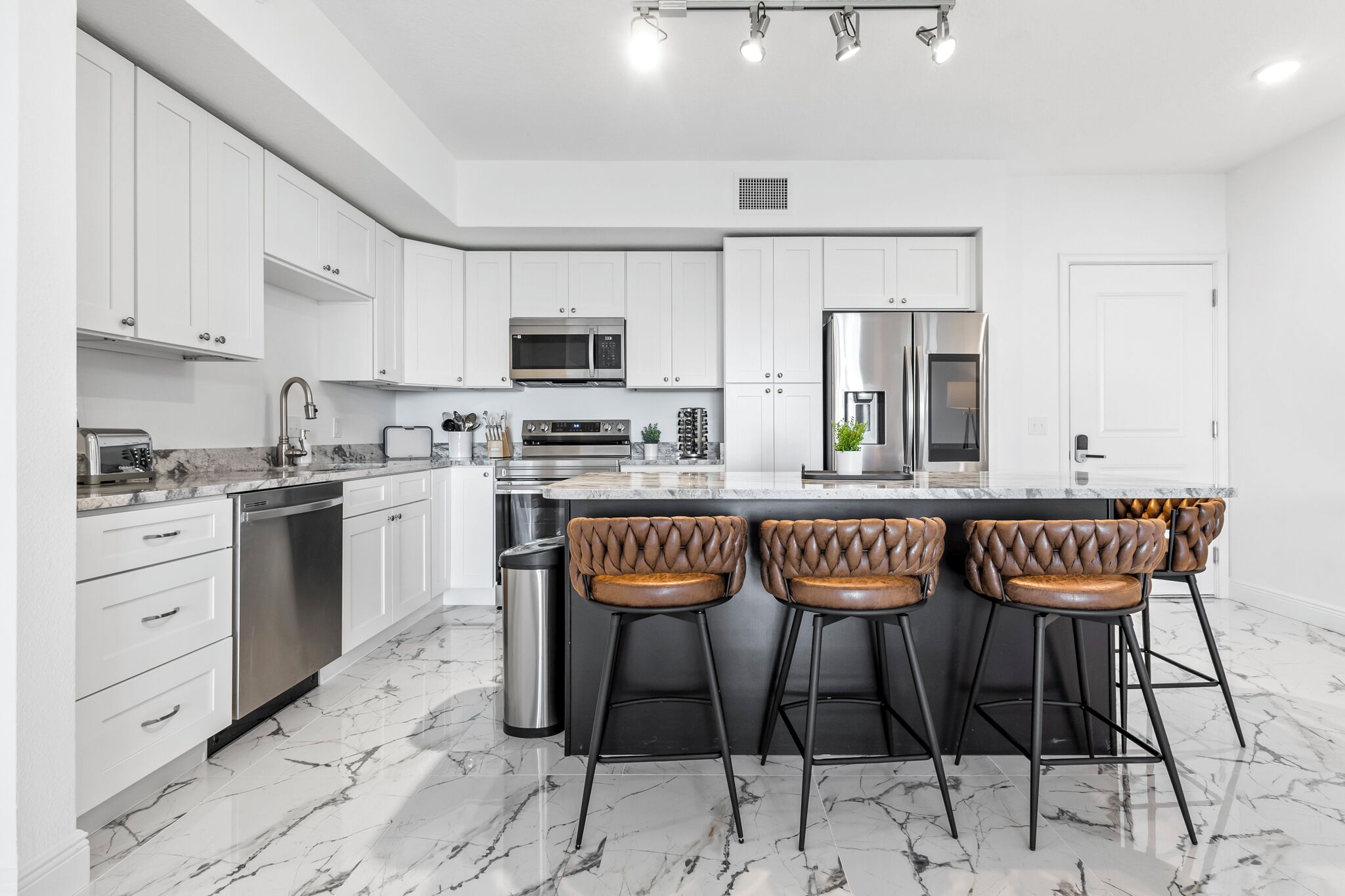 A modern kitchen with white cabinets, stainless steel appliances, and a marble floor. There are three bar stools with leather seats.