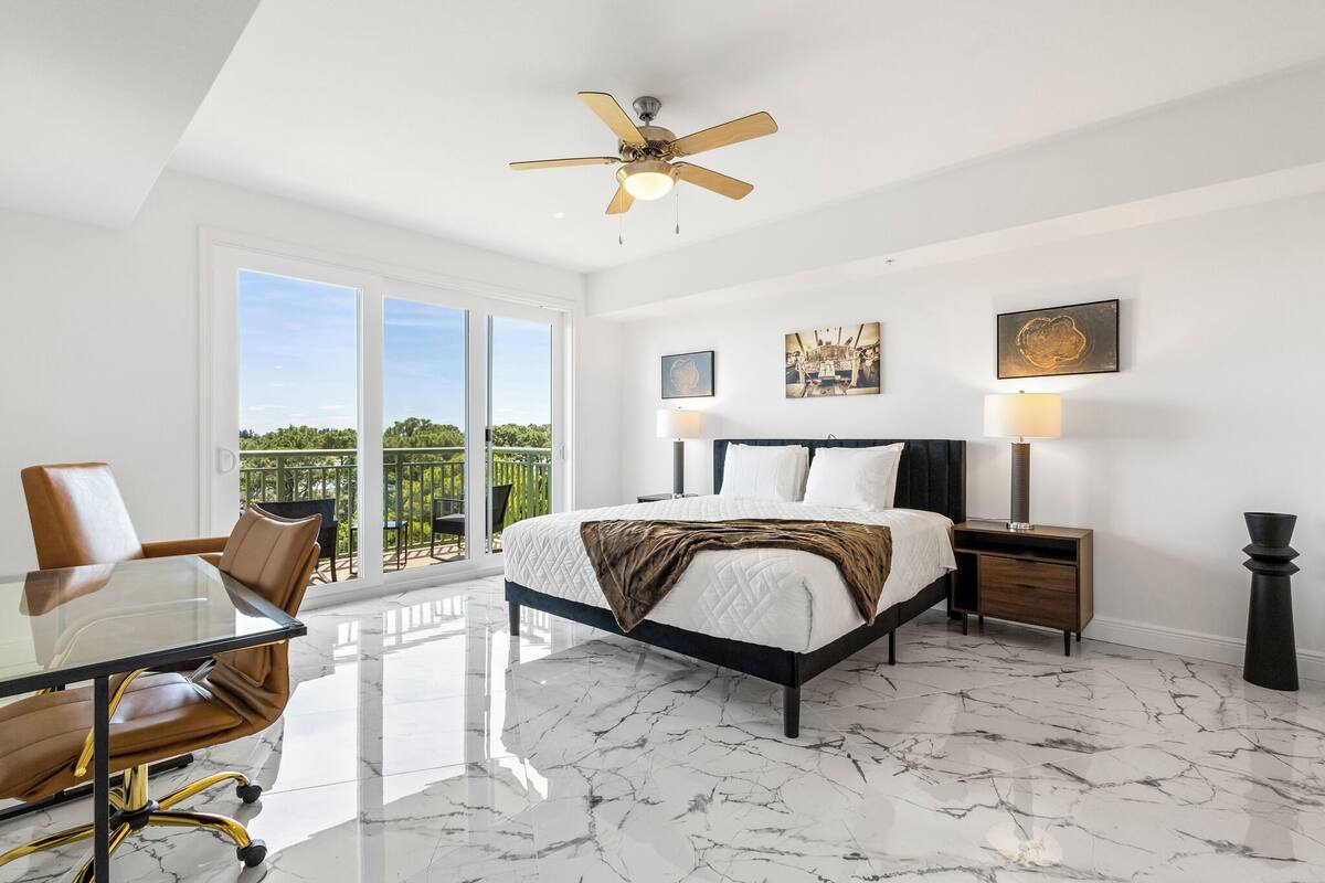 A bedroom with a bed, a desk, and a chair, a ceiling fan, and a balcony with a view.