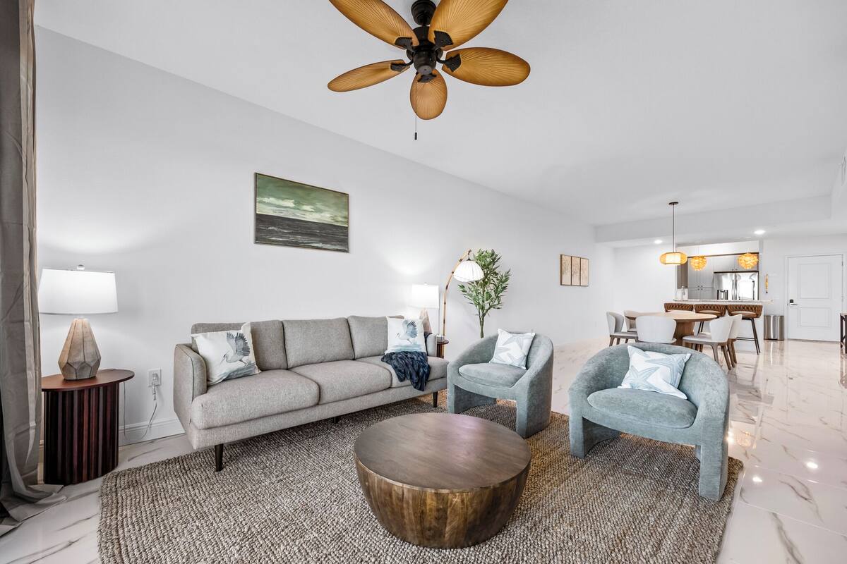 A modern living room with a sofa, two armchairs, a coffee table, a ceiling fan, a painting, and a dining area in the background.