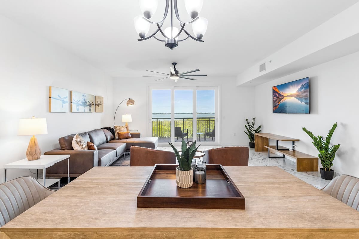 A modern living room with a dining table, sofa, and chairs. There is a ceiling fan, a chandelier, and a large window with a view of the outdoors.