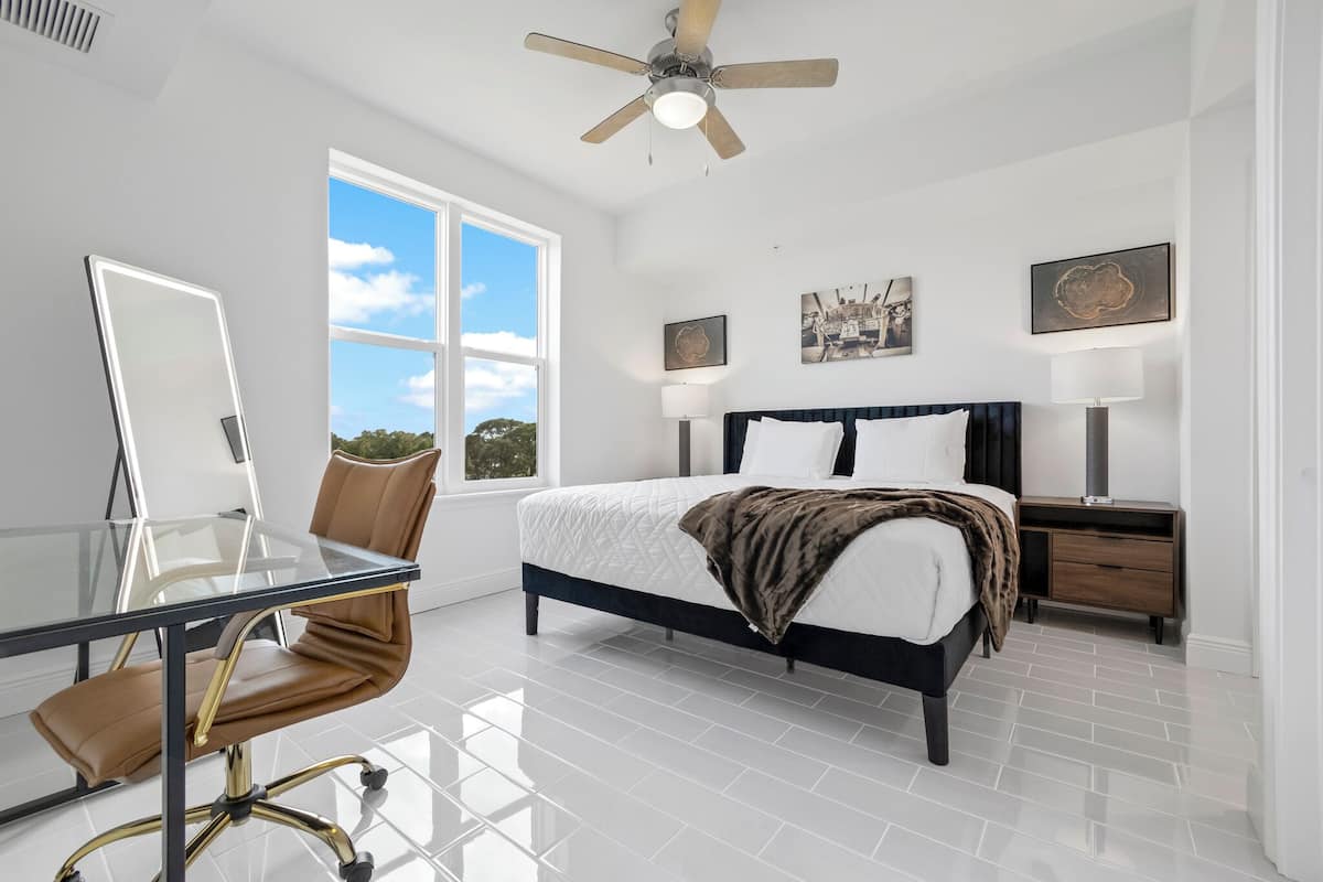 A bedroom with a bed, bedside tables, a desk with a chair, a ceiling fan, and a large window.