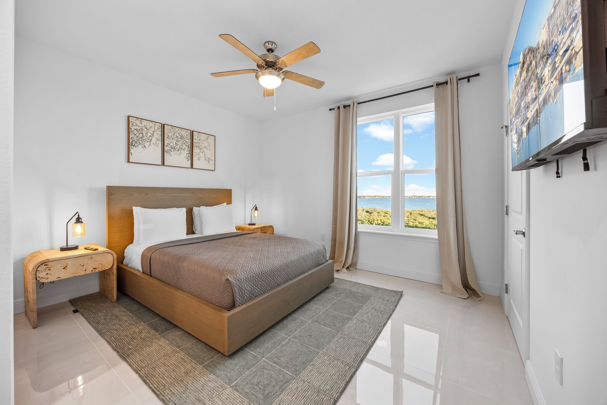 Une chambre moderne avec un lit en bois, un ventilateur de plafond, une grande fenêtre offrant une belle vue et un téléviseur à écran plat fixé au mur.