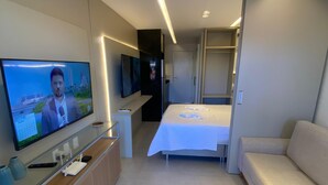 Een moderne hotelkamer met een bed, een slaapbank, een televisie op een standaard en een bureau met een computer en afstandsbedieningen.