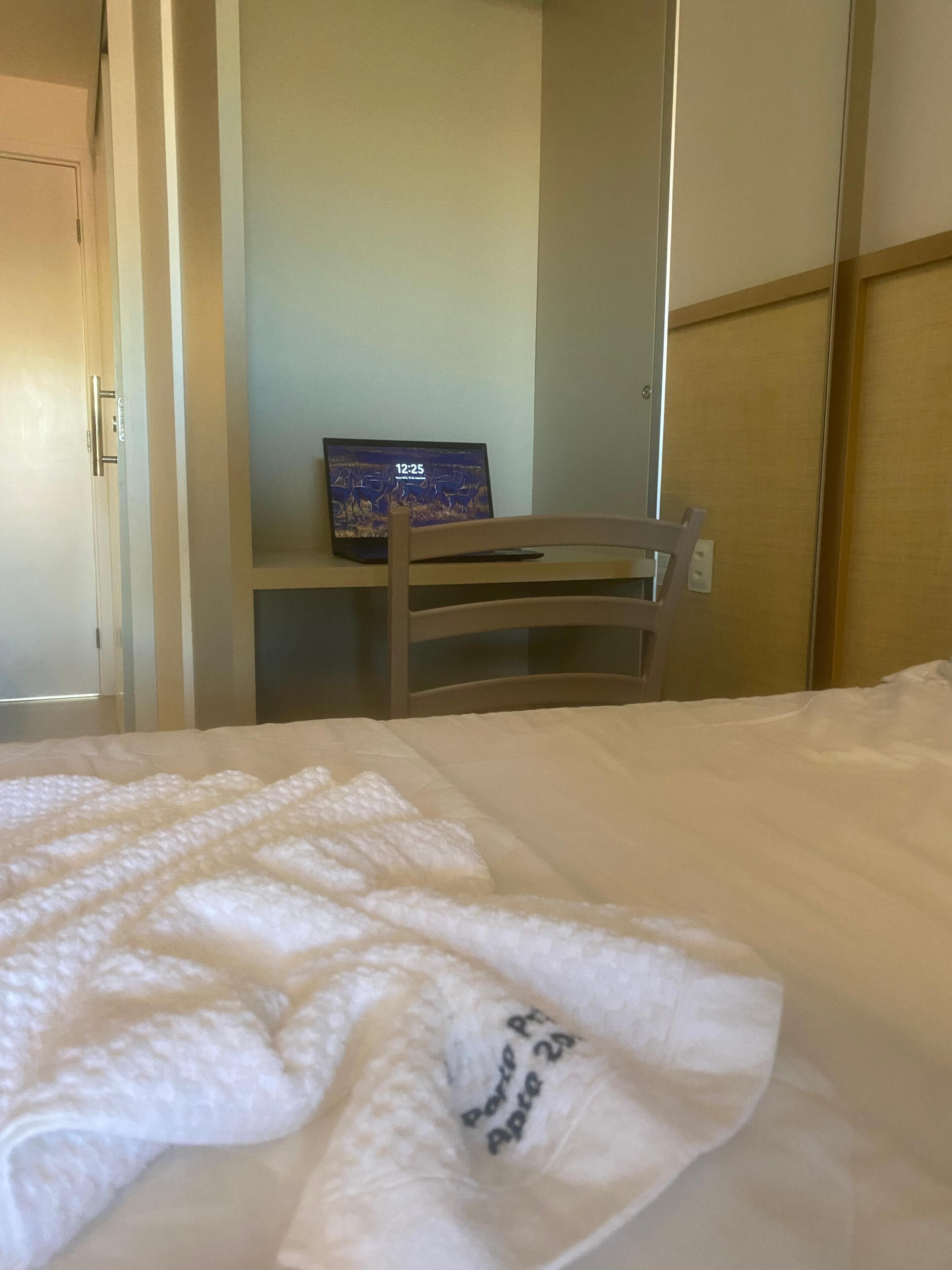 Una habitación de hotel con cama, una silla y un escritorio con una computadora portátil mostrando la hora.