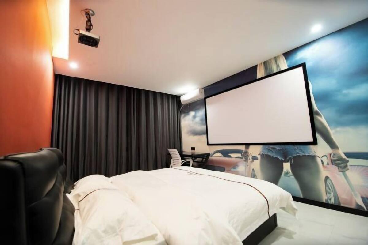 A hotel room with a bed, a projector screen, a desk with a chair, and a large painting on the wall.