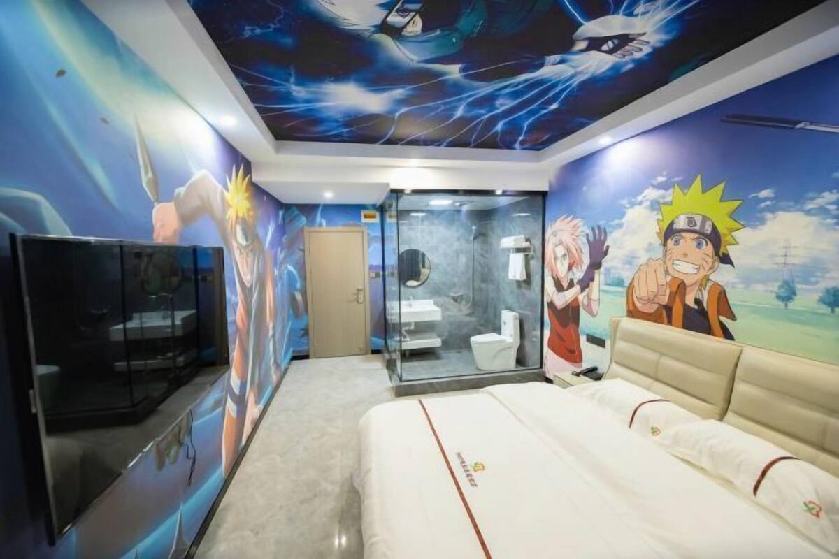 A hotel room with a bed, a TV, a bathroom with a glass partition, and anime-themed wall art.