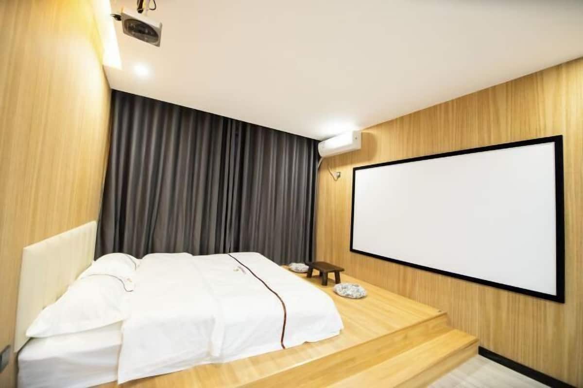 A hotel room with a bed, a large window, a projector screen, and a wooden wall.