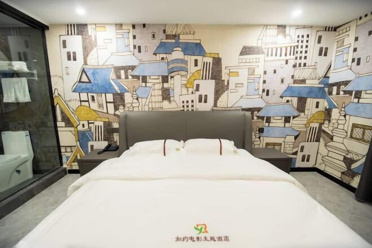 A hotel room with a bed, a nightstand, a wall with a cityscape mural, a window with a view, and a towel rack.