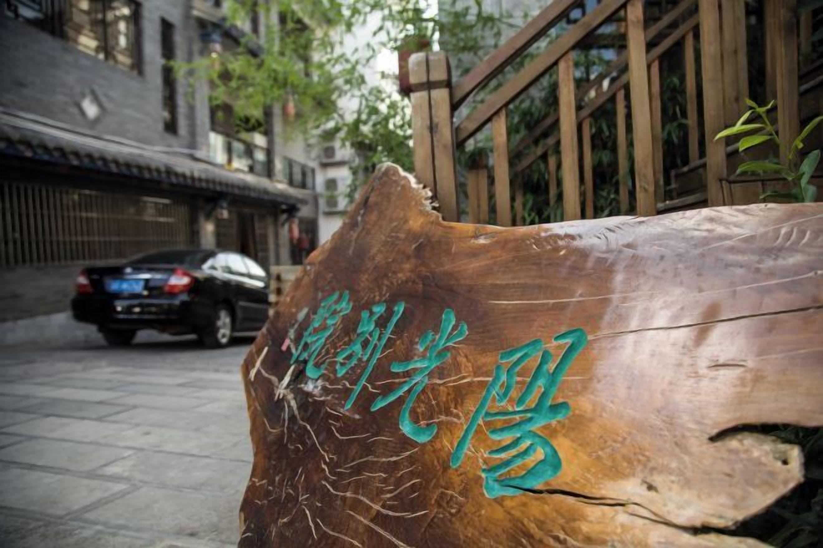 A wooden sign with carved text, a car parked on the street, and traditional buildings in the background.