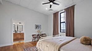 Ein Schlafzimmer mit einem großen Bett, einem Deckenventilator, einem Fenster mit Vorhängen, einem Schreibtisch mit Stuhl und Blick auf einen Küchenbereich.