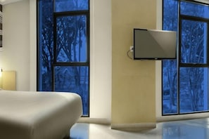 Ein Hotelzimmer mit Bett, einem flachbildfernseher an der Wand und großen Fenstern mit Blick auf Bäume.