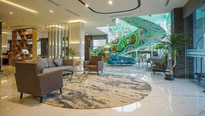 A spacious lobby with a modern design, featuring a large seating area with sofas and armchairs, a central coffee table, and a colorful mural on the wall. - Regantris Surabaya (Tegalsari)