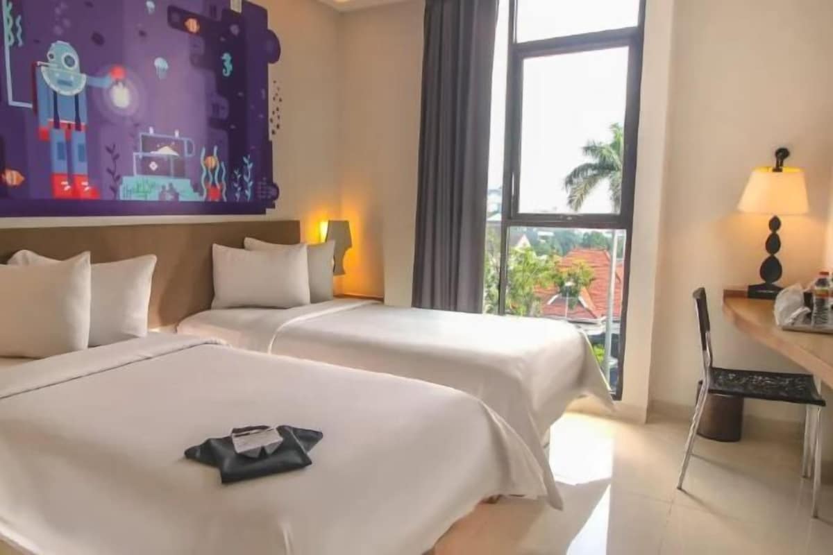 Ein Hotelzimmer mit zwei Betten, einem Schreibtisch mit Stuhl, einem Fenster mit Blick auf Palmen und einer Wand mit einem farbenfrohen Wandgemälde.